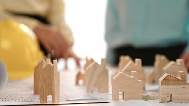 Architect and engineer discuss about building blueprint on table with architectural document, measurement tools scatter around at office, use wooden block for house positioning. Closeup. Delineation.