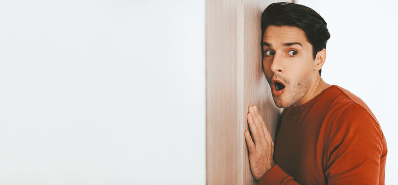 A young man in a red sweater expresses surprise and excitement while leaning against a light-colored wall. His facial expression conveys curiosity and anticipation in a contemporary indoor space.