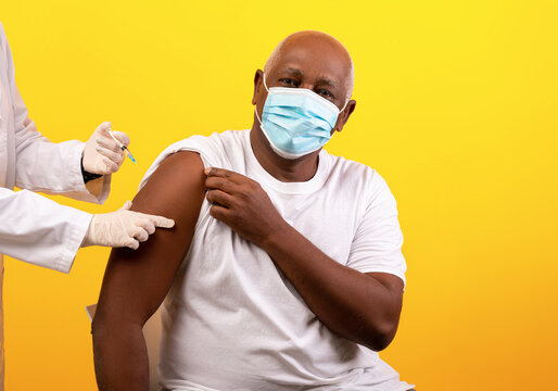 A senior African American man receives a coronavirus vaccine shot from a doctor. He wears a face mask and looks calm as he is being immunized against covid-19 in a bright studio.