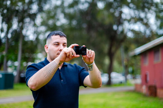 A man in his 30s stands in a park, taking photographs with a digital camera on an overcast day