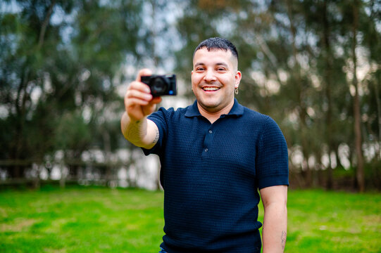 A man in his 30s happily takes a photo with his camera while standing in a grassy park.