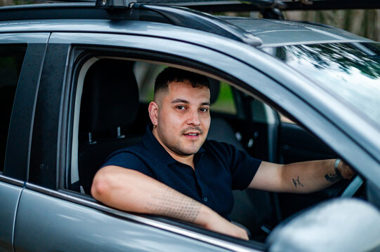 A man in his 30s sits comfortably in his car, smiling while parked in a park.