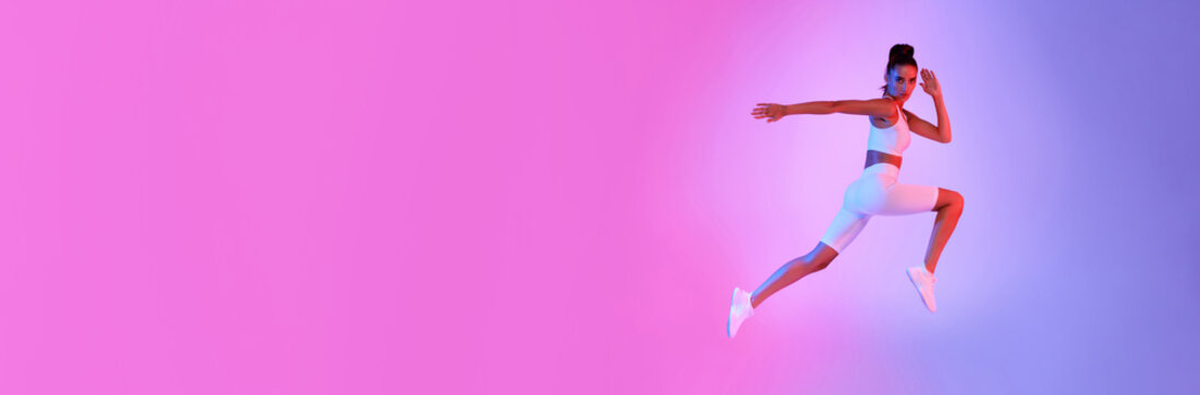 A sporty female is captured jumping in mid-air with her arms raised during a fitness workout. The vibrant pink and blue background enhances her energetic pose and showcases her athleticism perfectly.