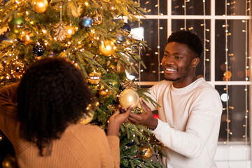 A happy young black couple happily decorates their Christmas tree with garlands and ornaments in a warm living room. They enjoy the festive season while preparing for New Year together.