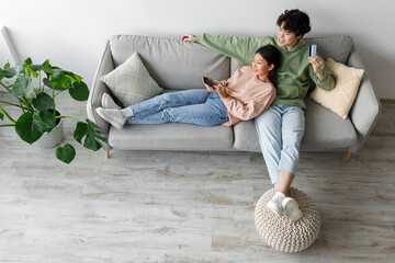 A happy Asian couple sits together on a comfortable couch at home, holding a smartphone and credit card while shopping online. They are relaxed and engaged in their activity.