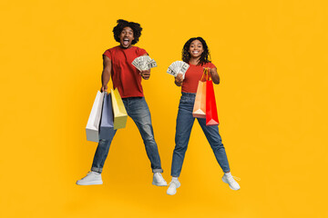 Fototapeta premium Two joyful friends jump against a bright yellow background, holding cash and colorful shopping bags. Their enthusiasm shows they are having a fun shopping day together.