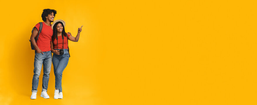 A young couple stands against a bright yellow backdrop, smiling and pointing. They are dressed casually in matching outfits, ready for an adventure with a camera in hand. - Powered by Adobe