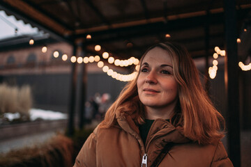 Warm portrait of a smiling woman in a puffer jacket under string lights at dusk, soft bokeh and cozy evening street atmosphere