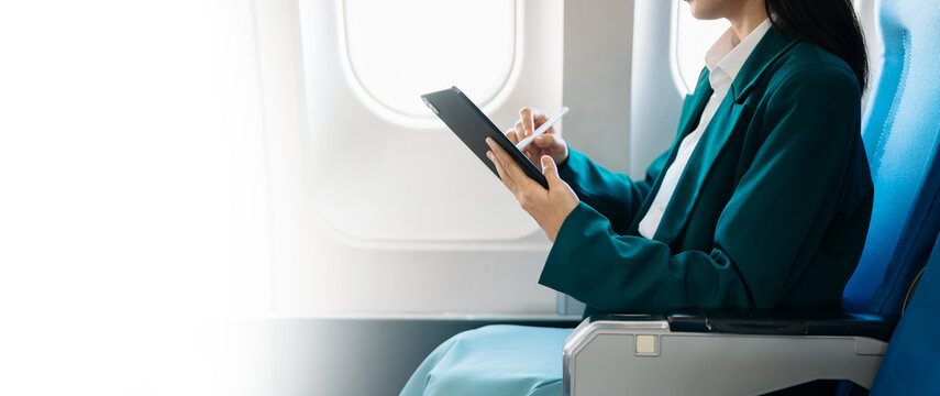 Asian businesswoman using tablet with stylus during airplane flight. Concept of business travel, digital lifestyle, and remote work flexibility.