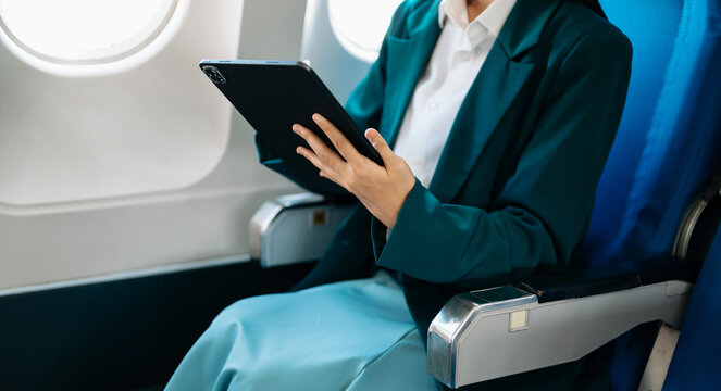 Asian businesswoman using tablet with stylus during airplane flight. Concept of business travel, digital lifestyle, and remote work flexibility.