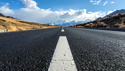 A black asphalt road stretches towards distant snow-capped mountains under a vast, partly cloudy blue sky, a picturesque landscape