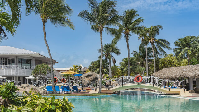 Swimming pool in a tropical resort. Arched pedestrian bridge over turquoise water. A lifebuoy on a rope railing. A canopy with a thatched roof. Palm trees against the blue sky. Cuba. Varadero. Hotel