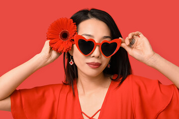 Pretty young Asian woman in stylish heart-shaped sunglasses with beautiful gerbera flower on red background