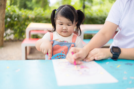 A cute little Asian girl sits at a table coloring with her mother teaching her next to her,Asian mothers teach their daughter to draw pictures. Concept of holiday activities.