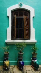 A traditional wooden window with shutters opens onto a vibrant teal wall at the Penang Peranakan Mansion. Below, three potted plants on elephant stands add cultural charm.