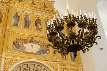 Large ornate bronze chandelier with candles illuminating a richly decorated gold-plated iconostasis inside an Orthodox church, featuring detailed religious paintings