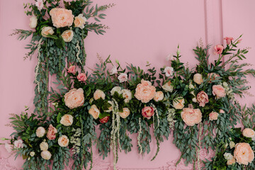 Lush pink and peach artificial roses, peonies, and green eucalyptus foliage creating a decorative floral arch or backdrop against a soft pastel pink wall
