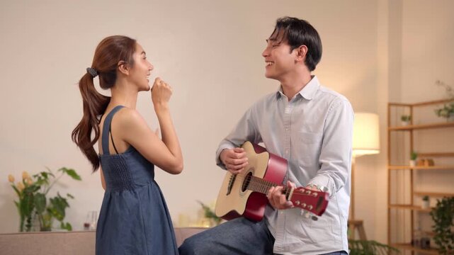 Asian young adult couple enjoy music together play acoustic guitar sing song home living room happy cheerful romantic moments indoors