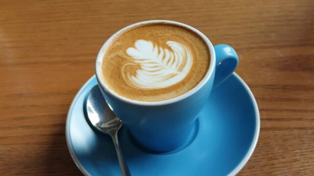 A close-up shot of a skilled barista creating beautiful latte art, specifically a heart design, with steamed milk into a blue ceramic coffee cup. The scene is set on a rustic wooden table, emphasizing