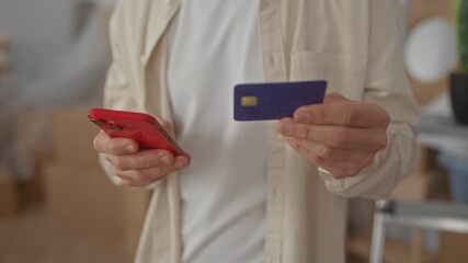 Caucasian man holding a smartphone and credit card in a new home's living room, indicating online shopping or payment activity in an indoor apartment setting. - Powered by Adobe