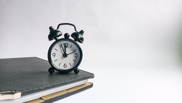 A small black alarm clock placed on top of a closed notebook over a white background, symbolizing time management, productivity, and organization in daily work or study.