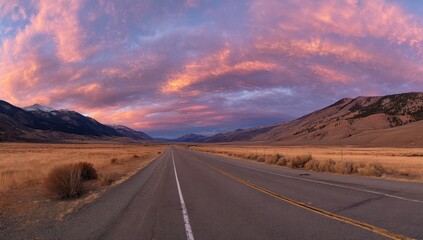 A wide, open road stretches through a valley bathed in the vibrant hues of a breathtaking sunset, framed by snow-capped mountains.