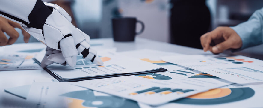 A robot hand collaborates with a human in a modern office, analyzing business reports and charts, showcasing the integration of technology in the workplace.Noogenesis - Powered by Adobe