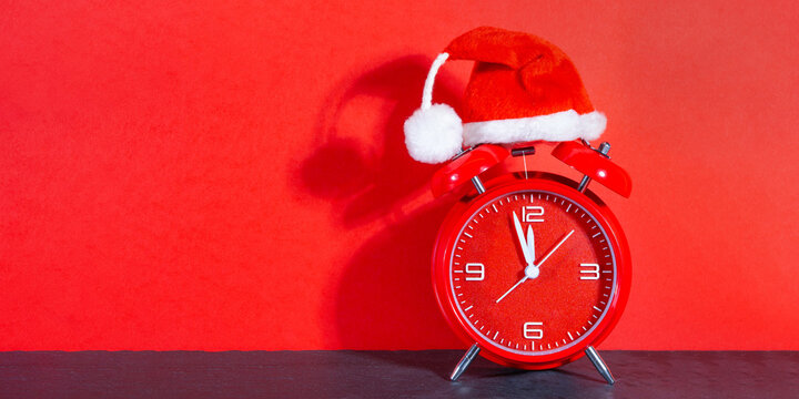 Christmas xmas saint nicholas day celebration holiday greeting card long - santa hat on red retro alarm clock on table, isolated on red background - Powered by Adobe