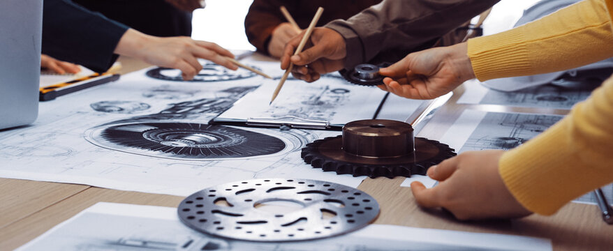 A diverse group engages in a collaborative design meeting, analyzing sketches and mechanical components on the table, promoting teamwork and innovation in engineering projects. SACTR