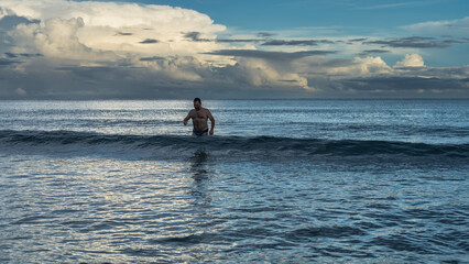 Early morning. A man in swimming trunks stands in the ocean wave. Glare on the surface of the water. The clouds in the blue sky are illuminated with golden light. Malaysia. Borneo.  Kota Kinabalu.