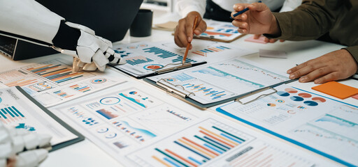 A modern workspace showcases a collaborative meeting with human and robot hands analyzing various business reports, charts, and data on a table.Noogenesis