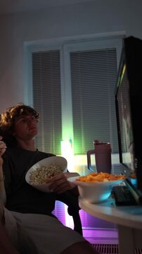 Vertical screen young student talks while watching evening content eating popcorn in a neon lit room creating a relaxed casual atmosphere that reflects late night habits and typical carefree student m