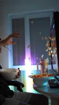 Vertical screen young man throws popcorn and chips at monitor showing frustration and disappointment with video during night entertainment surrounded by neon lights and snacks creating chaotic atmosph