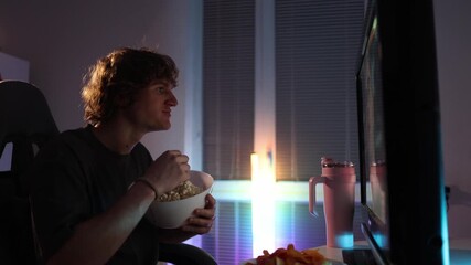 Young man watches screen at night holding popcorn focused and absorbed calm room atmosphere shows dependency on endless content and casual snacking as part of his daily routine without noticing time p