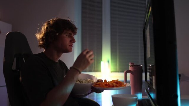 Young man sits close to screen eating popcorn and chips at night ignoring health and focusing on constant video consumption casual room with neon light atmosphere of unhealthy habit moment