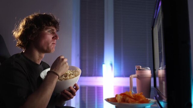 Young man sits close to screen eating popcorn and chips at night showing dependence on endless online content and unhealthy food habits quiet room neon light mood of passive consumption