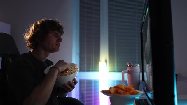 Student watches the screen at night instead of studying eating popcorn and chips neon light fills the room creating a quiet relatable moment of procrastination and low motivation at home