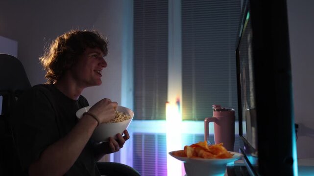 Young man absentmindedly watches the screen while eating popcorn late at night showing a mindless addicted state neon light fills the room capturing a simple yet relatable moment of scrolling without 