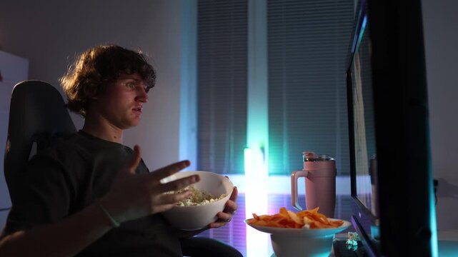 Young man frustrated by boring content throws popcorn at the monitor at night no longer feeling entertained neon light fills the room creating a chaotic impulsive moment of irritation