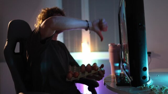 Young man gives thumbs down to the monitor holding a carton of eggs at night neon light fills the room showing frustration and disappointment before impulsive action in a tense moment