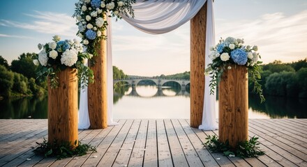 A beautiful wedding archway adorned with white and blue flowers stands on a wooden deck, overlooking a serene river and a distant bridge at sunset, creating a romantic scene
