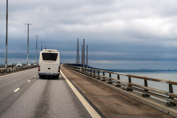 White campervan traveling on the Oresund Bridge, connecting Denmark viewable to the city skyline in Sweden under cloudy sky. 