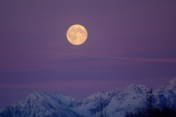 The full moon rises over Alaska's snowy Talkeetna Range at sunset.