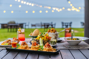 Outdoor dining setup with gourmet food, tropical cocktails, and ocean backdrop. Perfect for elegant events or beach restaurants. Decorative lighting enhances atmosphere.