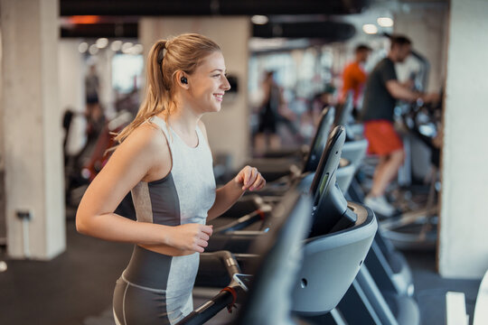 Smiling young woman running on treadmill indoors in modern gym environment with fitness equipment