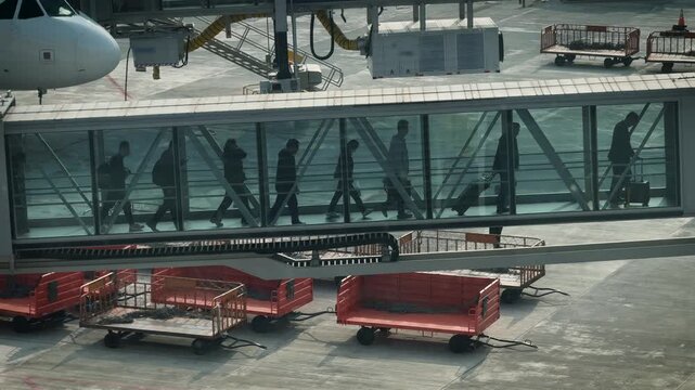 Passengers walk through an airport jet bridge after arriving on a commercial flight. Silhouettes of travelers with carry-on bags move toward the terminal, seen through the glass walkway, slowmotion.