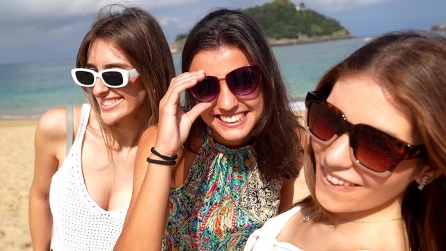 Three beautiful young women with sunglasses smiling and posing for a selfie on a sunny beach, having fun together during their summer vacation and enjoying the beautiful seaside scenery - Powered by Adobe