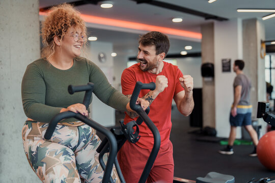 Smiling woman exercises on stationary bike while motivated man cheers in indoor gym workout - Powered by Adobe