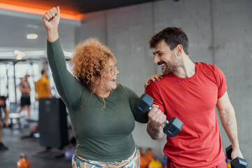 Middle aged woman and young man exercising together in indoor gym smiling and lifting dumbbells