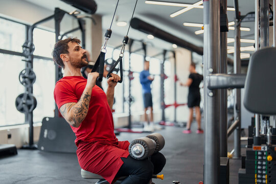 Focused young man in red exercising with pulley machine in modern gym with others working out